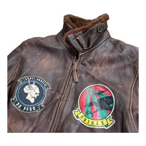 Op de afbeelding: Bruine leren bomberjack met een schapenvachtkraag en meerdere patches. De patches bevatten een schedelontwerp en het woord "AVIREX". De jas heeft een ritssluiting en een verweerde uitstraling.