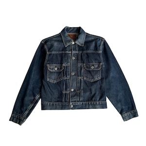 May include: A dark blue denim jacket featuring a classic collar, long sleeves with buttoned cuffs, and a front button closure. The jacket includes two chest pockets with buttoned flaps and vertical stitched pleats running down the front panel.
