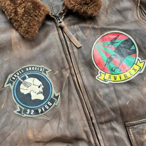 Op de afbeelding: Een bruine leren jas met een bruine bontkraag en een ritssluiting. De jas heeft twee patches: een met een schedel en de woorden "Skull Angels" en "32 PFSQ", en een andere met een vogelafbeelding en het woord "Avirex".