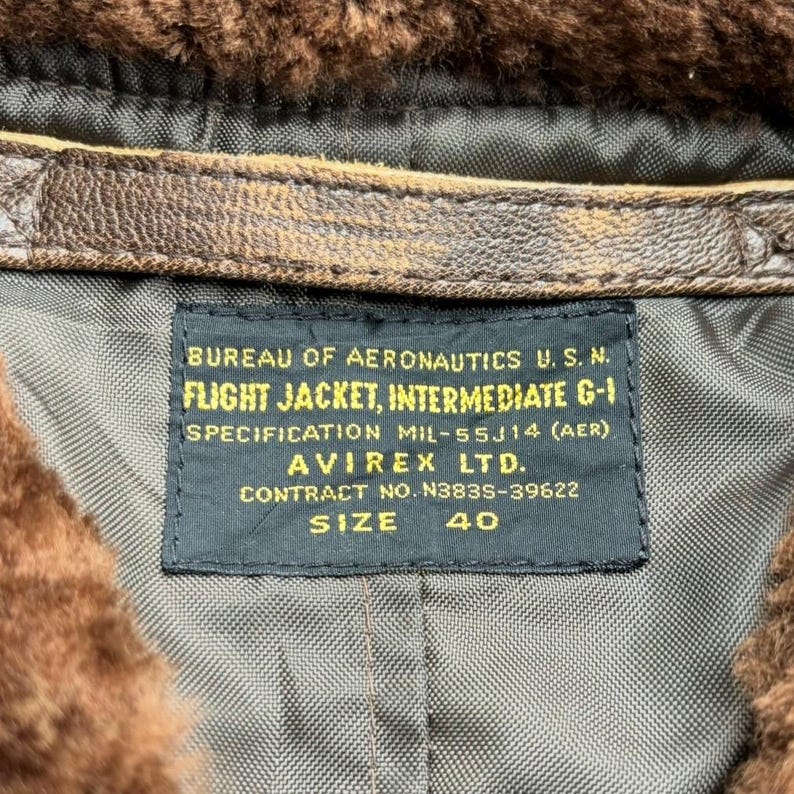 Op de afbeelding: Close-up van een vintage flight jacket label. Het label luidt "BUREAU OF AERONAUTICS U.S.N. FLIGHT JACKET, INTERMEDIATE G-1." Het label bevat ook de specificatie, fabrikant, contractnummer en maat 40.