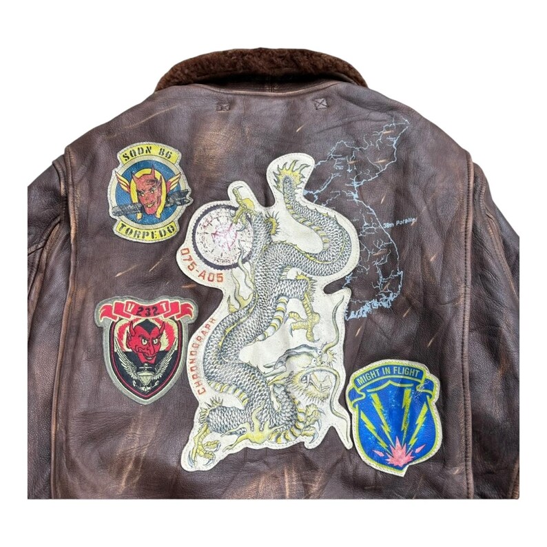 Op de afbeelding: Bruine leren bomberjack met meerdere patches. Patches tonen een duivel, een draak en een kaart. De jas heeft een bruine bontkraag en een versleten uitstraling. Met tekst zoals "Torpedo".