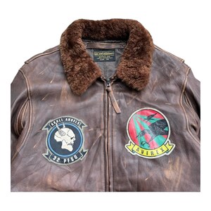 Op de afbeelding: Bruine leren bomberjack met een bruine bontkraag. De jas heeft twee patches: een met een schedel en de tekst "SKULL ANGELS 32 PFSQ", en een andere met een vogelafbeelding en de tekst "AVIREX".