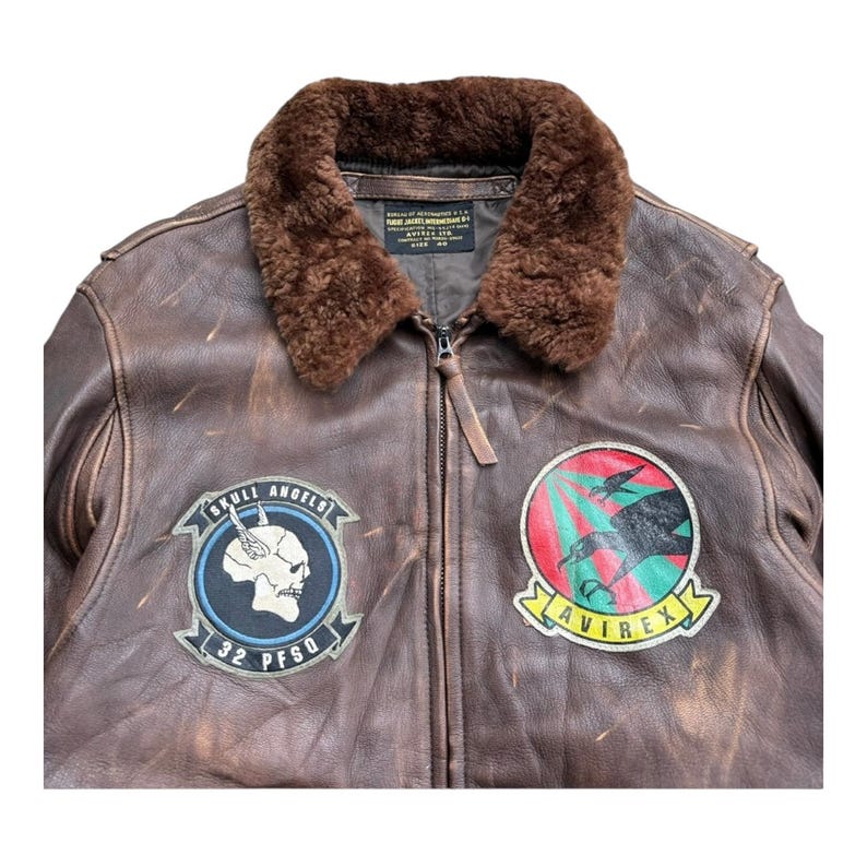 Op de afbeelding: Bruine leren bomberjack met een bruine bontkraag. De jas heeft twee patches: een met een schedel en de tekst "SKULL ANGELS 32 PFSQ", en een andere met een vogelafbeelding en de tekst "AVIREX".