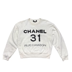 May include: Cream-coloured sweatshirt with long sleeves. The front displays the text "CHANEL 31 RUE CAMBON" in black, sequined lettering. The number "31" is prominently displayed below the brand name.