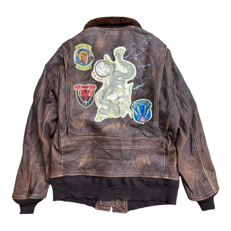 Op de afbeelding: Bruine leren bomberjack met patches. De jas heeft een bruine bontkraag, zwarte geribde zoom en manchetten, en geborduurde patches met militaire ontwerpen. De jas heeft een versleten, vintage uitstraling.