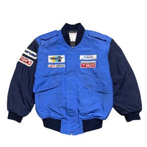May include: A blue and navy racing jacket featuring various patches, such as "TRUST", "APEX", "Netz", and "TRD". The jacket has a zip-up front, two front pockets, and ribbed cuffs and hem. The sleeves are navy, and the body is blue.