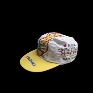 May include: White and yellow baseball cap with the word "SUZUKI" on the brim. The cap features a graphic of a musician playing a guitar with flames and the text "Rock 'N' Roll Marathon San Diego."