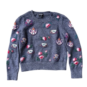 Könnte beinhalten: Ein grauer Pullover mit gestickten Blumen-, Teetassen- und Teekannen-Designs. Der Pullover hat einen Rundhalsausschnitt und lange Ärmel. Die Stickereien umfassen rosa, rote, weiße und grüne Akzente. Der Markenname "CHANEL" ist sichtbar.