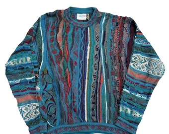 May include: A colorful, textured sweater with a crew neck. The sweater features a variety of patterns and colors, including teal, blue, red, and white. The brand name "GOGI" is visible on the neckline. The sweater has long sleeves and a ribbed hem.
