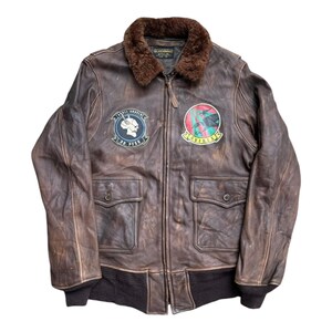 Op de afbeelding: Bruine leren bomberjack met een bruine bontkraag. De jas heeft twee voorzakken, een geribbelde zoom en patches op de borst. E&eacute;n patch heeft een schedel en de tekst "32 PIGE". De andere patch heeft een rood en groen ontwerp met de tekst "AVIREX".