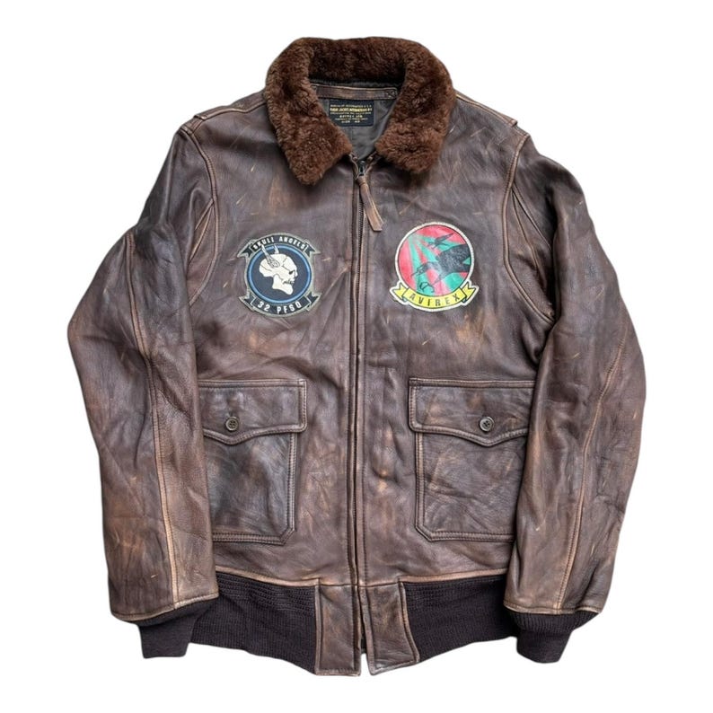 Op de afbeelding: Bruine leren bomberjack met een bruine bontkraag. De jas heeft twee voorzakken, een geribbelde zoom en patches op de borst. E&eacute;n patch heeft een schedel en de tekst "32 PIGE". De andere patch heeft een rood en groen ontwerp met de tekst "AVIREX".