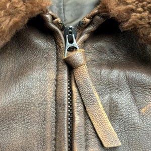 Op de afbeelding: Close-up van een bruine leren jas met een rits. De jas heeft een schapenvachtkraag en een leren ritstrekker. De rits is zilverkleurig en heeft de merknaam "AVIREX".