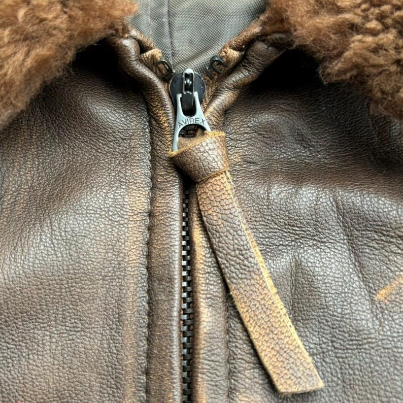 Op de afbeelding: Close-up van een bruine leren jas met een rits. De jas heeft een schapenvachtkraag en een leren ritstrekker. De rits is zilverkleurig en heeft de merknaam "AVIREX".