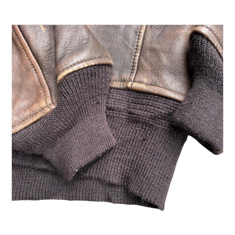 Op de afbeelding: Close-up van een bruine leren jas met donkerbruine gebreide manchetten en tailleband. Het leer heeft een versleten uiterlijk met zichtbare stiksels en een vintage esthetiek. De manchetten en tailleband zijn geribbeld.