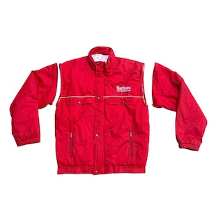 May include: A red jacket with detachable sleeves. The jacket features a stand-up collar, two front pockets with zippers, and a white trim. The Marlboro logo is printed on the upper left chest area. The jacket is made of a lightweight material.