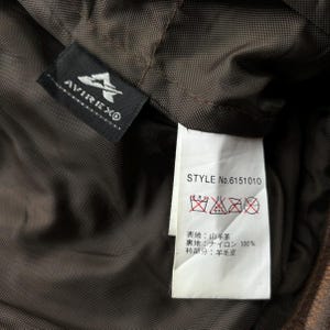 Op de afbeelding: Close-up van de binnenkant van een bruine jas, met een zwart label met het "AVIREX&reg;"-logo en een wit label met wasvoorschriften en het stijlnummer "No. 6151010". De voering van de jas is donkerbruin.