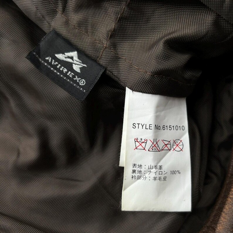 Op de afbeelding: Close-up van de binnenkant van een bruine jas, met een zwart label met het "AVIREX&reg;"-logo en een wit label met wasvoorschriften en het stijlnummer "No. 6151010". De voering van de jas is donkerbruin.