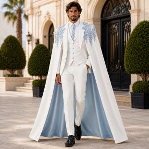 May include: A white suit featuring a matching cape with light blue floral embroidery on the shoulders. The ensemble includes a white vest with light blue buttons, a white dress shirt, a light blue tie, and white trousers with a light blue lining.