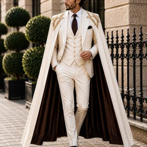 May include: A cream-colored three-piece suit consisting of a vest, trousers, and a blazer, paired with a white dress shirt and a dark brown tie. A long, floor-length cream cape with gold embroidery on the shoulders and a dark brown lining is worn.