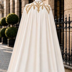 May include: A floor-length off-white cape featuring intricate gold embroidery across the upper back and shoulders. The garment has a structured collar and a full, flowing silhouette that drapes down to the ground, creating a clean and elegant appearance.