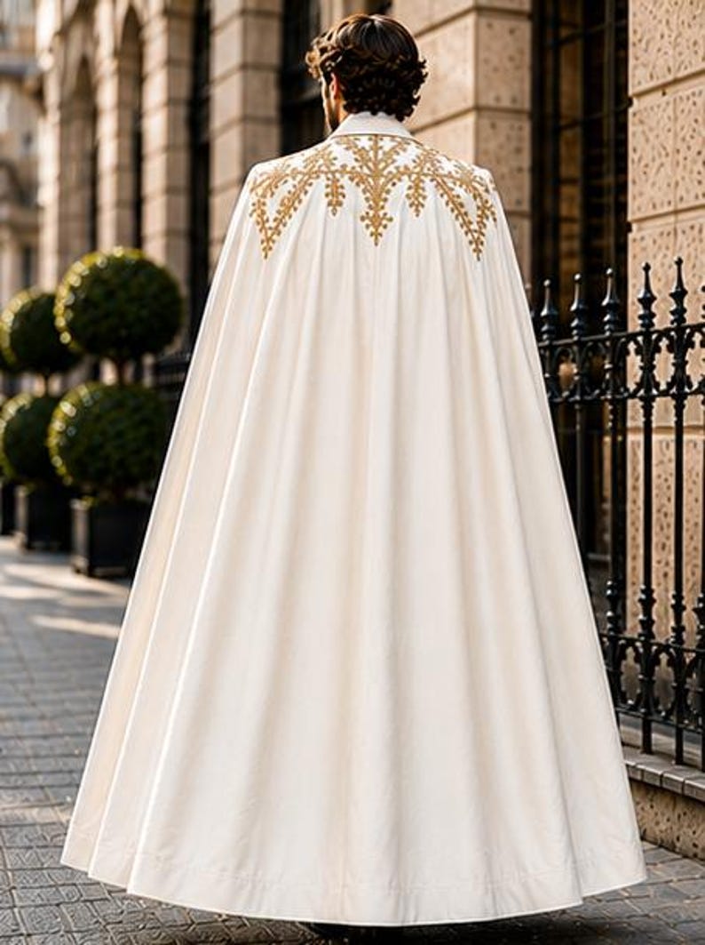May include: A floor-length off-white cape featuring intricate gold embroidery across the upper back and shoulders. The garment has a structured collar and a full, flowing silhouette that drapes down to the ground, creating a clean and elegant appearance.
