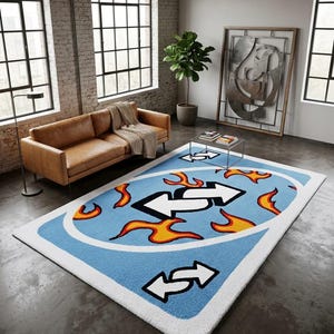 May include: A large, rectangular rug with a light blue background and a white border. The rug features a design of white arrows and orange flames. The rug is placed on a concrete floor in a modern living room setting.