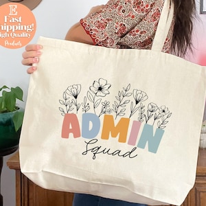 May include: Cream-colored tote bag with the words "ADMIN Squad" in colorful block letters, with floral illustrations above. The bag is held by a person. The bag has a simple design, perfect for carrying items.