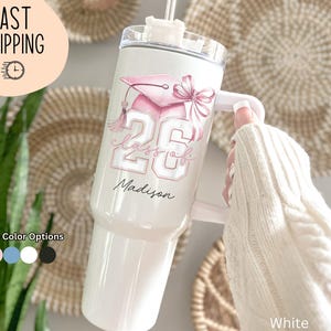 May include: White tumbler with a pink graduation cap and bow design. The tumbler features the text "Class of 26" and the name "Madison". The tumbler has a handle and a straw. Additional color options are available.