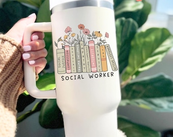 May include: A cream-colored tumbler with a white handle and lid, featuring a floral design above a stack of books labeled with social work values. The text "SOCIAL WORKER" is printed below the books. A white straw extends from the lid.
