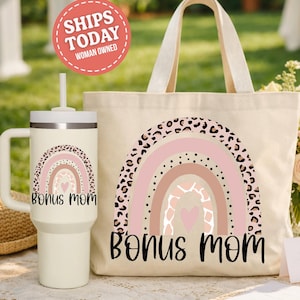 May include: A cream-colored tumbler with a handle and straw, and a canvas tote bag, both featuring a rainbow design with leopard print accents and the words "Bonus Mom" in black script. The tote bag has a cream-colored handle.
