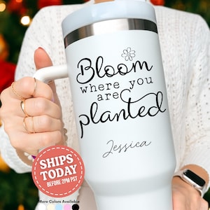 May include: White insulated tumbler with a handle, featuring the text "Bloom where you are planted" in black script, along with a small flower graphic. The name "Jessica" is written below the quote. The tumbler has a light blue lid.