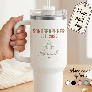 May include: A white stainless steel tumbler with a handle and a clear lid featuring a straw. The text reads SONOGRAPHER EST. 2025 with a graphic of an ultrasound probe and the name Hannah written in script below the graphic with a small heart.