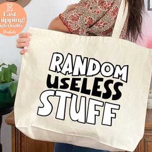 May include: Cream-colored tote bag with the text "RANDOM USELESS STUFF" in a bold, black and white design. The bag is held by a person. The background includes a lava lamp and a plant.