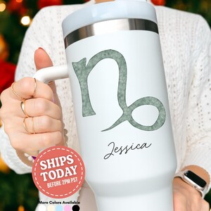 May include: White insulated tumbler with a light gray Capricorn zodiac symbol and the name "Jessica" in black script. The tumbler has a white handle and lid, and a silver band. The tumbler is designed to hold approximately 30 ounces.
