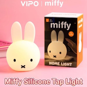 Könnte beinhalten: Ein weißes Miffy-Hasen-Nachtlicht aus Silikon mit schwarzen Augen und einem X-förmigen Mund. Daneben steht eine schwarz-orangefarbene Produktverpackung mit der Aufschrift Miffy Home Light, einer Abbildung der Lampe und dem Text VIPO | miffy.