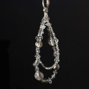 May include: A clear, teardrop-shaped beaded ornament. The ornament features a silver-colored loop at the top and is strung with clear, faceted beads and larger, round, clear beads with a gold shimmer. The background is black.