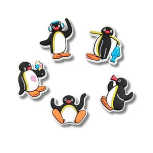 Op de afbeelding: Een set van vijf schoenbedels met het personage Pingu in verschillende houdingen, waaronder een met een blauwe koptelefoon, een met een blauwe vis, een met een ijsje, een zittend en een staand met een open snavel, in zwart, wit en oranje.