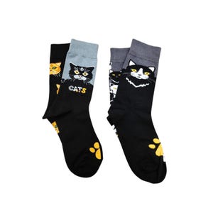 May include: Two pairs of black socks with cat-themed designs. One pair features a black cat face with the word "CATS" and paw prints. The other pair has a white and black cat face and paw prints. The socks have gray and black cuffs.
