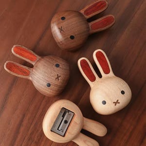 May include: Four wooden rabbit-shaped pencil sharpeners. Three are shown from the front with dark wood or light wood finishes, black eyes, and an X-shaped mouth. The ears have red wood inlays. One light wood sharpener is shown from the back.