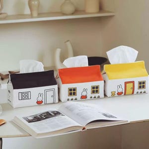 Peut inclure: Trois housses de boîte à mouchoirs en tissu rectangulaires en forme de petites maisons. L'une est noire et blanche, l'autre orange et blanche, et la troisième jaune et blanche. Chacune présente un lapin, des fenêtres et une porte imprimés.