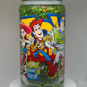 May include: A clear glass tumbler filled with green glitter and liquid, featuring a colorful Toy Story graphic with Woody, Buzz Lightyear, Jessie, and an alien. The lid is topped with clear, ice-like acrylic pieces, and the cup has a smooth, cylindrical shape.