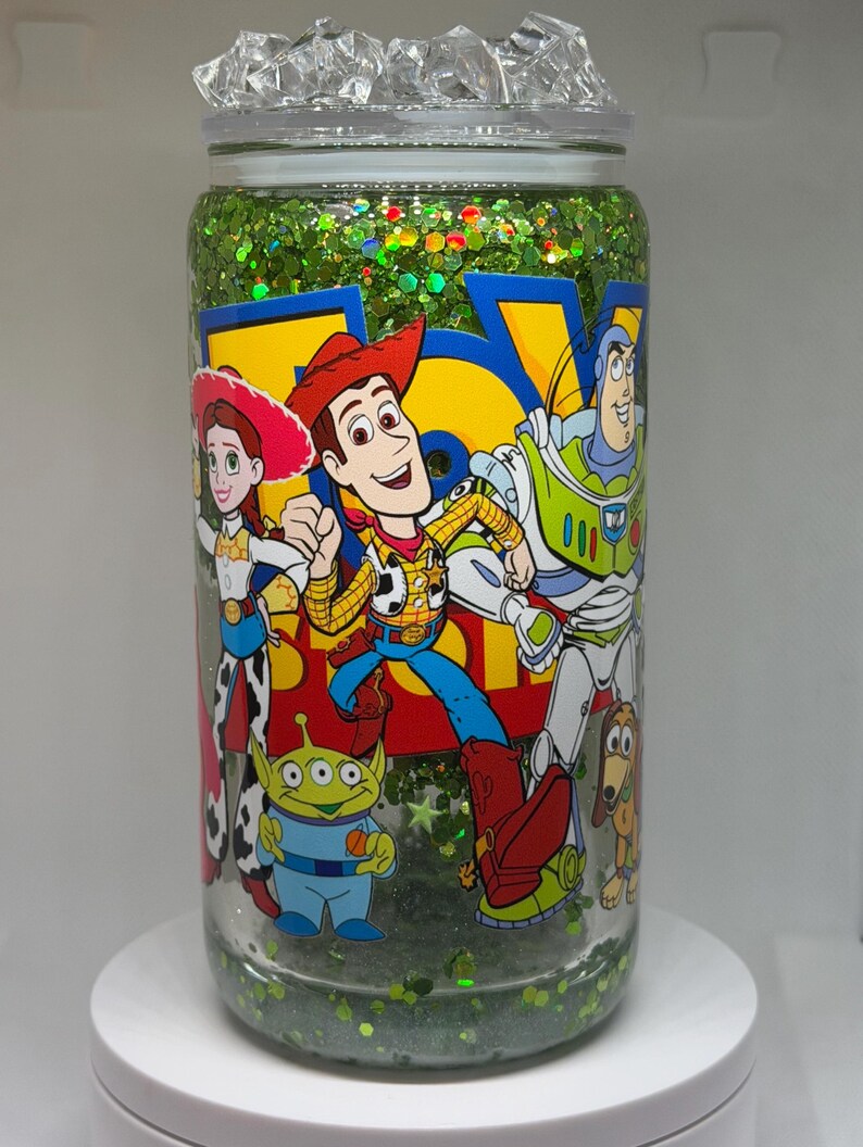 May include: A clear glass tumbler filled with green glitter and liquid, featuring a colorful Toy Story graphic with Woody, Buzz Lightyear, Jessie, and an alien. The lid is topped with clear, ice-like acrylic pieces, and the cup has a smooth, cylindrical shape.