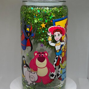 May include: A clear glass tumbler filled with green glitter and liquid, featuring a wrap-around graphic of various animated characters including a pink bear, a cowgirl, a penguin, a pig, and a villain. The lid is topped with clear crystals.