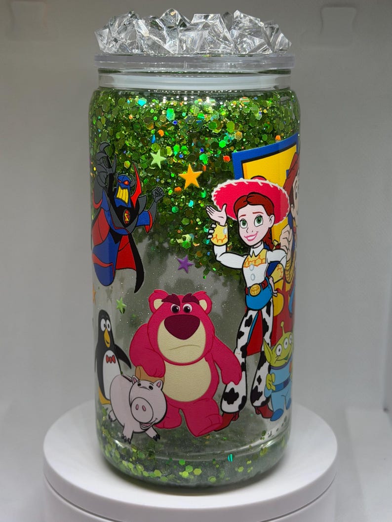 May include: A clear glass tumbler filled with green glitter and liquid, featuring a wrap-around graphic of various animated characters including a pink bear, a cowgirl, a penguin, a pig, and a villain. The lid is topped with clear crystals.