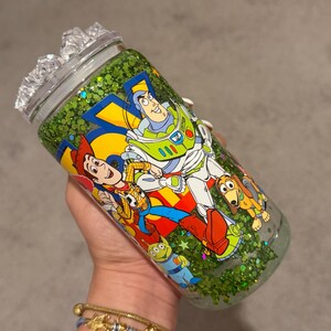 May include: A clear glass tumbler with a lid, filled with green glitter. The exterior features a colorful graphic print of Woody, Buzz Lightyear, Slinky Dog, and an Alien from Toy Story. The design includes bold yellow and blue accents.