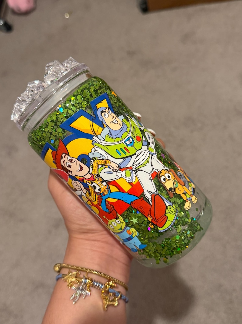 May include: A clear glass tumbler with a lid, filled with green glitter. The exterior features a colorful graphic print of Woody, Buzz Lightyear, Slinky Dog, and an Alien from Toy Story. The design includes bold yellow and blue accents.