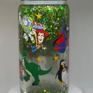 May include: A clear glass tumbler filled with green glitter and small yellow star shapes. The exterior features colorful cartoon character decals including a space ranger, a cowboy, a green dinosaur, and a penguin. The lid is topped with clear ice-like pieces.