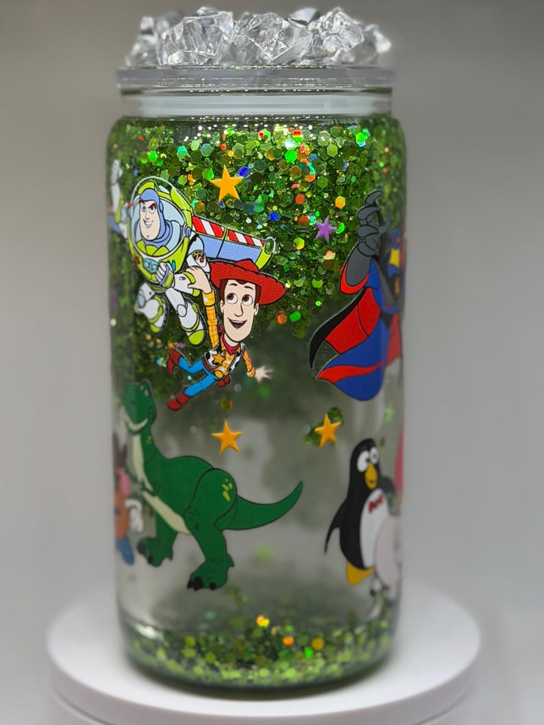 May include: A clear glass tumbler filled with green glitter and small yellow star shapes. The exterior features colorful cartoon character decals including a space ranger, a cowboy, a green dinosaur, and a penguin. The lid is topped with clear ice-like pieces.