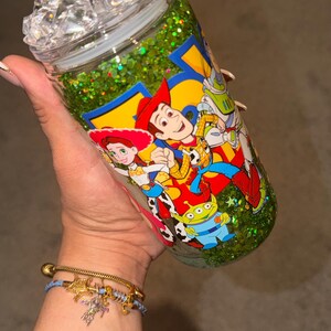 May include: A clear glass tumbler filled with green glitter liquid, featuring a Toy Story character graphic with Woody, Jessie, and an alien. The lid is topped with clear, faceted, ice-like plastic cubes, and the cup has a smooth, cylindrical shape.