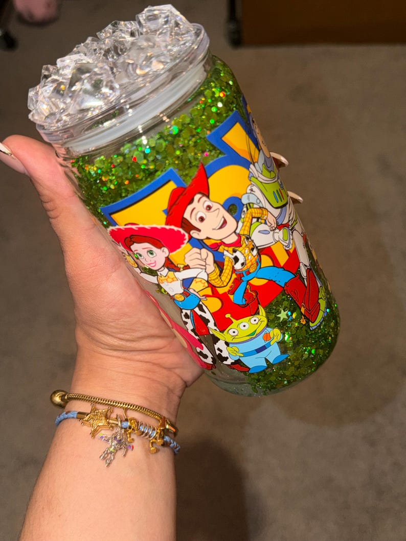 May include: A clear glass tumbler filled with green glitter liquid, featuring a Toy Story character graphic with Woody, Jessie, and an alien. The lid is topped with clear, faceted, ice-like plastic cubes, and the cup has a smooth, cylindrical shape.